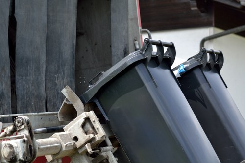 Close-up view of two black rubbish bins with closed lids, attached to a metal waste collection vehicle, in an urban environment. The bins are made of plastic with a slightly textured surface and are positioned side by side, mounted securely to the vehicle's compartment. Visible metal handles are on the sides of each bin for lifting, and the bins are slightly tilted to fit into the vehicle's loading mechanism. The background includes a wooden fence or wall with dark horizontal panels, and part of the vehicle's metallic framework is visible on the left, featuring mechanisms for securing and lifting the bins. The scene suggests a waste collection process, possibly in a city area near Fitzrovia, with natural daylight illuminating the scene, highlighting the clean and well-maintained condition of the bins and equipment used by Fitzrovia House Clearance for rubbish removal services.