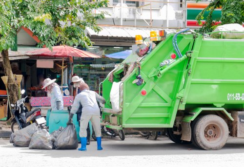 A bright green rubbish collection truck parked on a street in Fitzrovia, with two waste operatives actively collecting and loading black garbage bags from the sidewalk into the rear of the vehicle. The scene is set amidst urban surroundings, including a shopfront with a small awning and a tree with green foliage providing some shade. One worker is bending forward, holding a bag, while the other stands nearby, both wearing protective gloves and uniforms suitable for waste collection. In the background, there are additional street objects such as a motorcycle and various street furniture. The truck's surface appears metallic with visible wheels and mechanical components, and the setting is illuminated by natural daylight, emphasizing the urban environment typical of central London boroughs like Fitzrovia. This scene exemplifies routine rubbish removal services offered by Fitzrovia House Clearance in the local area, with a focus on efficient waste handling on urban streets.