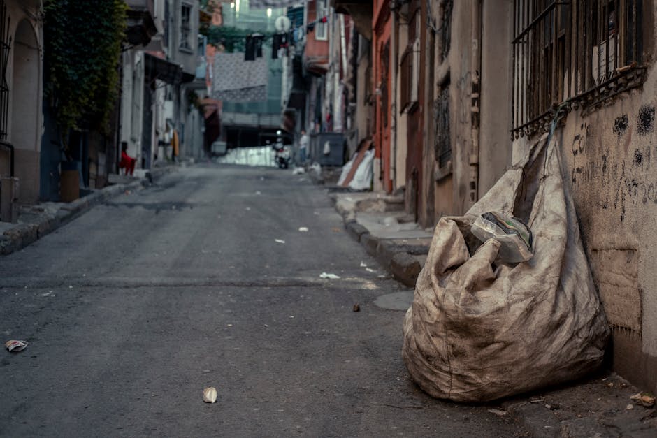 A street scene featuring a white waste collection truck with an open rear compartment actively engaged in rubbish removal. The truck's metallic surface shows signs of wear and rust around the edges of