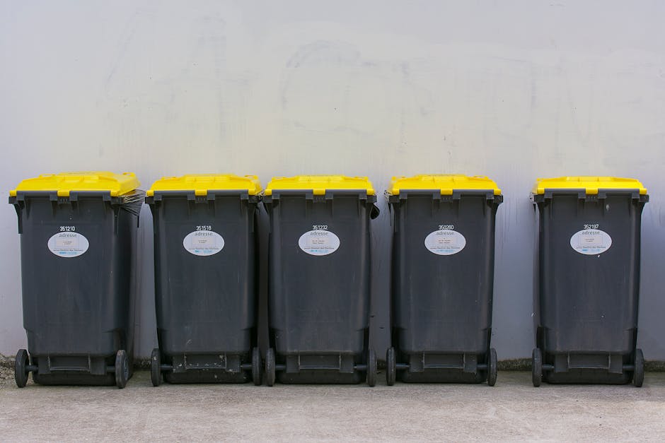 A large green waste bin filled with various discarded items, situated outdoors on a paved surface, likely in an urban environment nearby a residential or commercial area. The bin contains a mix of mat