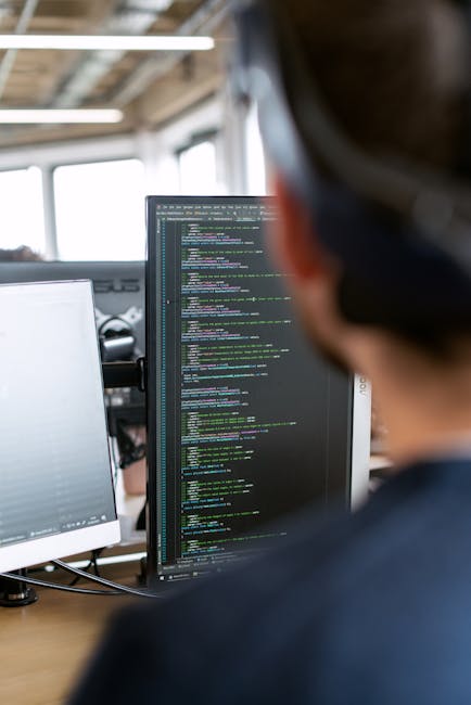 A person with short brown hair, wearing glasses, is sitting at a desk in a modern office environment, closely examining computer screens displaying lines of code with multicolored syntax highlighting.