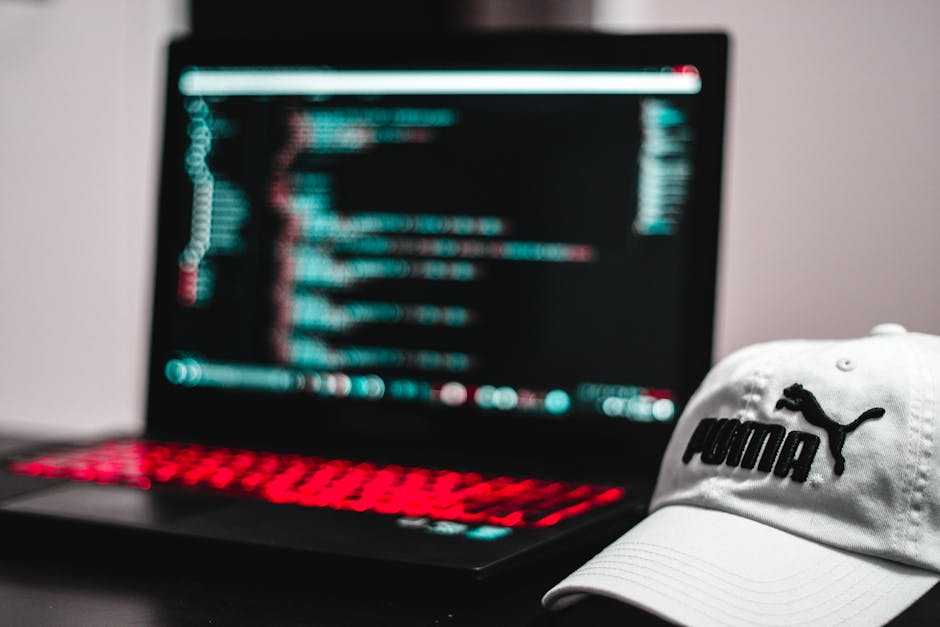 A person with short brown hair, wearing glasses, is sitting at a desk in a modern office environment, closely examining computer screens displaying lines of code with multicolored syntax highlighting.
